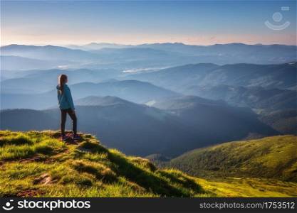 Girl on mountain peak with green grass looking at beautiful mountain valley in fog at sunset in summer. Landscape with sporty young woman, foggy hills, forest, sky. Travel and tourism. Hiking