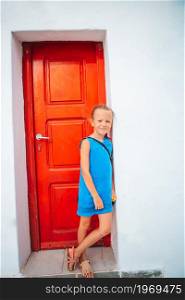 Girl on Mykonos vacation outdoors near red tradition greek door. Adorable little girl at old street of typical greek traditional village