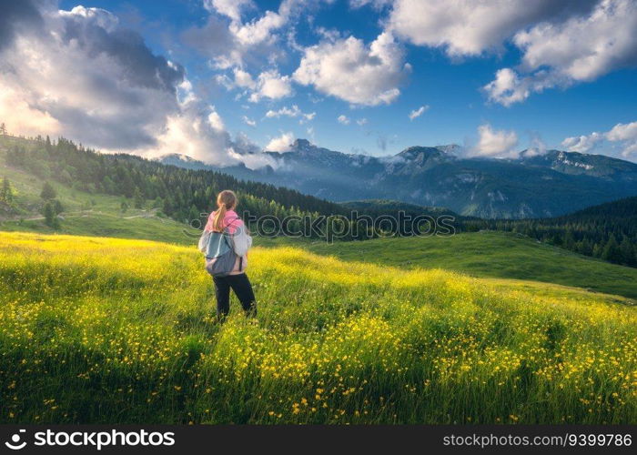 Girl on the hill with yellow flowers and green grass in beautiful alpine mountain valley at sunset in summer. Landscape with young woman in alps, trees, sky with clouds. Travel and Hiking. Slovenia