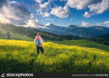 Girl on the hill with yellow flowers and green grass in beautiful alpine mountain valley at sunset in summer. Landscape with young woman in alps, trees, sky with clouds. Travel and Hiking. Slovenia