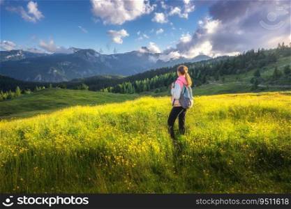 Girl on the hill with yellow flowers and green grass in beautiful alpine mountain valley at sunset in summer. Landscape with young woman in alps, rees, sky with clouds. Travel and Hiking. Slovenia