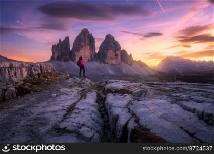Girl on the mountain peak and high rocks at colorful sunset in autumn. Tre Cime, Dolomites, Italy. Beautiful landscape with young woman on trail, cliffs, purple sky with pink clouds in fall. Hiking. Girl on the mountain peak and high rocks at colorful sunset