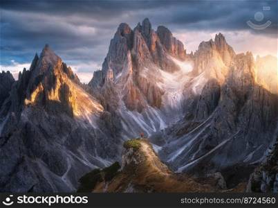 Girl on the mountain peak and high rocks at colorful sunset in autumn. Tre Cime, Dolomites, Italy. Colorful landscape with woman on trail, cliffs, grass, cloudy sky in fall. Hiking. Aerial view. Girl on the mountain peak and amazing high rocks at sunset