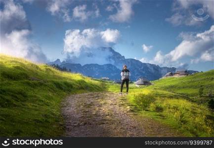 Girl on the rural road with yellow flowers and green grass in beautiful alpine mountain valley at sunset in summer. Young woman in old alpine village, sky with clouds. Travel and Hiking. Slovenia