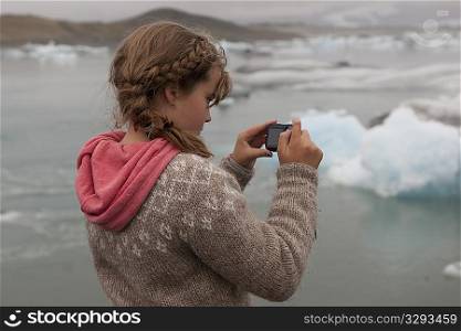 Girl photographer in an Icelandic sweater and a coral hoodie focussing on blue icebergs in a glacial lake