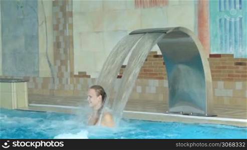 Girl playing in a swimming pool standing under a jet of water from a curved poolside water feature