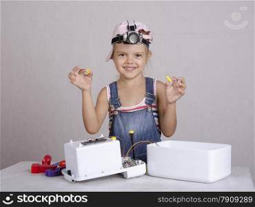 Girl playing in the repairer and repairs toy microwave. In one hand it a cog in another nut. Girl with fun and pleasure looks into the frame, and raised her hands. The child&#39;s head wearing a flashlight