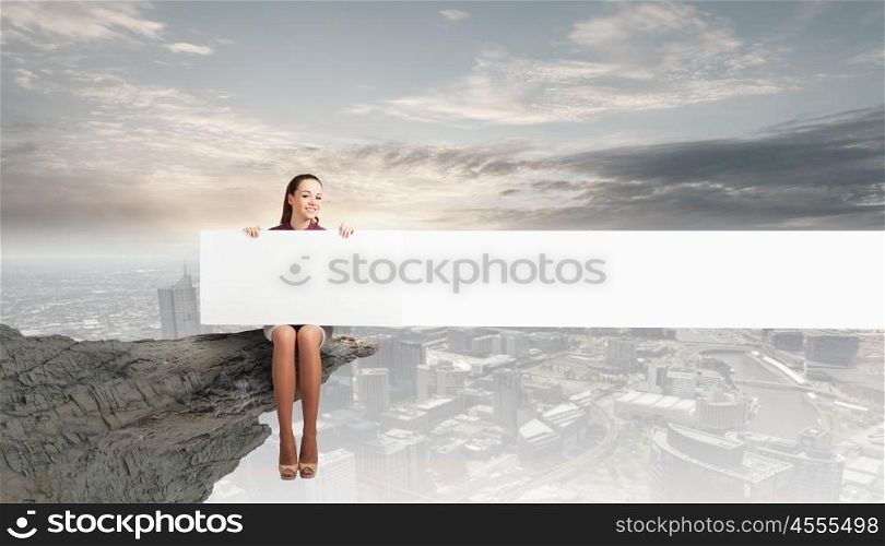 Girl presenting something. Young woman with white blank banner sitting on rock top