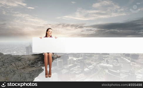 Girl presenting something. Young woman with white blank banner sitting on rock top