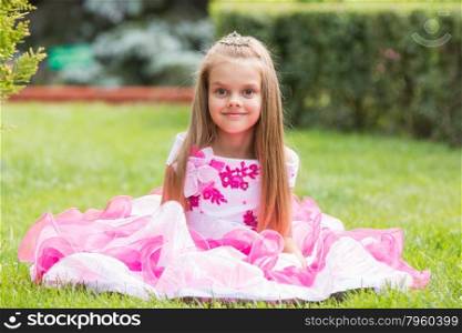 Girl princess sat on the lawn in the green garden. Happy six year old girl in a lush evening pink dress walking through the green garden
