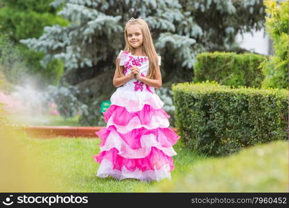 Girl princess stands on the lawn in the green garden. Happy six year old girl in a lush evening pink dress walking through the green garden