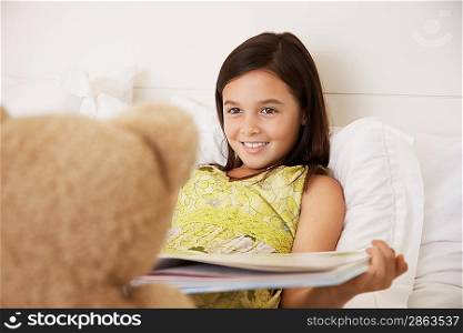 Girl reading story to teddy bear sitting on bed