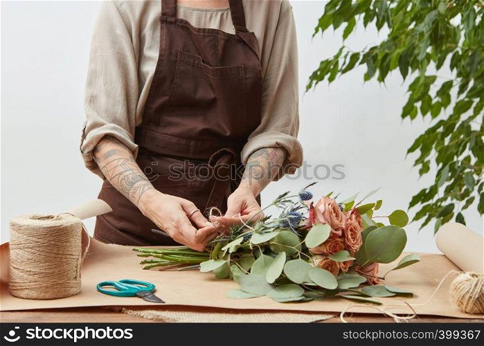 Girl's florist hands with tatoo are making bouquet with fresh flowers roses living coral color at the table with paper and rope. Process step by step. Concept floral shop.. Florist woman is creating fresh rose bouquet on a gray background. Process step by step. Small business with flowers delivery. Mother's Day.