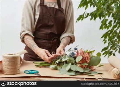 Girl's florist hands with tatoo are making bouquet with fresh flowers roses living coral color at the table with paper and rope. Process step by step. Concept floral shop.. Florist woman is creating fresh rose bouquet on a gray background. Process step by step. Small business with flowers delivery. Mother's Day.