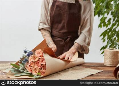 Girl's florist hands with tatoo are making bouquet with fresh flowers roses living coral color at the table with paper and rope. Process step by step. Concept floral shop.. Florist woman is wrapping fresh rose bouquet in a decorative paper on a wooden table on a light wall background. Process step by step. Mother's Day holiday.