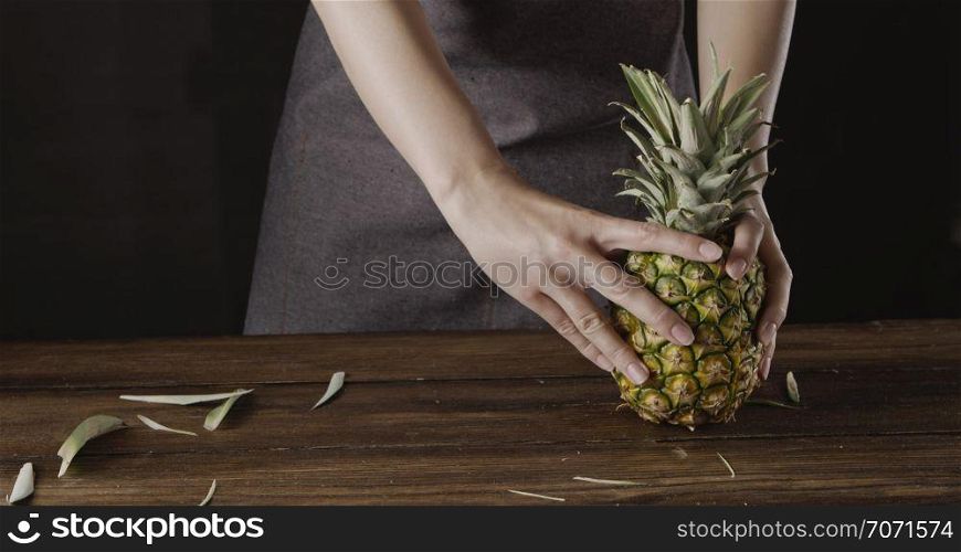 Girl&rsquo;s in a textile apron holds fresh ripe juicy natural organic texoticl fruit pineapple on a wooden kitchen table on a black background. Concept of vegetarian diet eating.. Woman&rsquo; hands with ripe juicy natural organic tropical fruit pineapple on a wooden kitchen table on a black background.