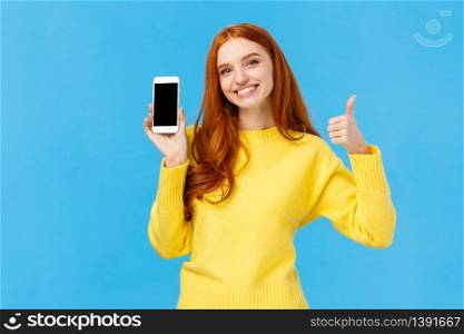 Girl showing good app, recommend download very useful application. Cute redhead woman in yellow sweater, like winter holiday sale in online store, showing smartphone display, blue background.. Girl showing good app, recommend download very useful application. Cute redhead woman in yellow sweater, like winter holiday sale in online store, showing smartphone display, blue background