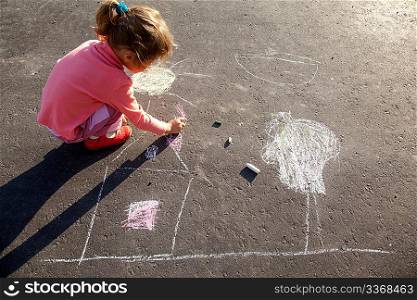 girl sits on concrete asphalt square road. girl draws painting line sun house tree a chalk on asphalt. chld drawings paintings on asphalt concrete. back side view