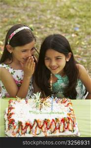 Girl sitting in front of a birthday cake and whispering to her friend
