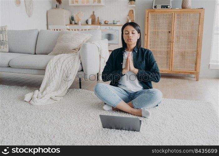 Girl sitting in front of laptop in lotus pose and meditating. Young hispanic woman is practicing yoga at home. Posture exercises, relaxation, stress relief. Online learning, internet fitness training.. Girl sitting in front of laptop in lotus pose and practicing yoga at home. Posture exercises.