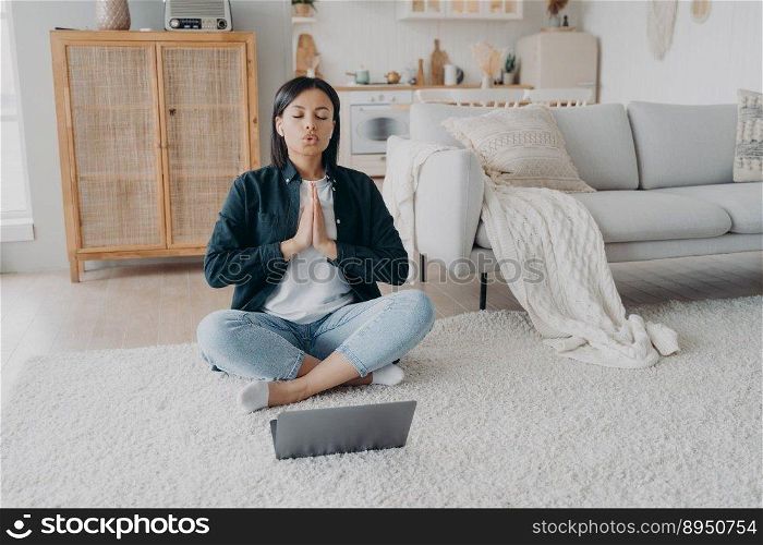 Girl sitting in front of laptop in lotus pose and meditating. Young hispanic woman is practicing yoga at home. Posture exercises, relaxation, stress relief. Online learning, internet fitness training.. Girl sitting in front of laptop in lotus pose and practicing yoga at home. Posture exercises.