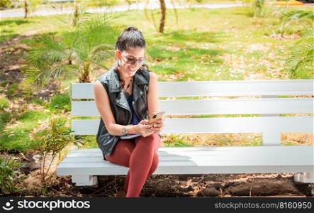 Girl sitting on a bench checking her cell phone, Happy woman sitting in a park texting on her cell phone, Woman sitting on a bench sending a text message