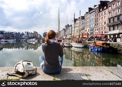 girl sitting on a pier and taking a shot of Honfleur harbor with her smartphone