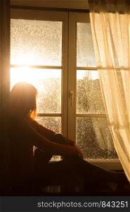 girl sitting on a windowsill and looking at the evening city at the beams of orange sunset sun