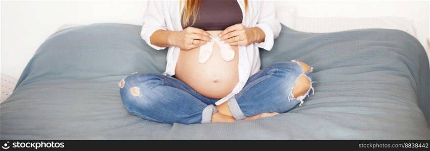 girl sitting on bed in bedroom and holding pink socks near her pregnant belly.. girl sitting on bed in bedroom and holding pink socks near her pregnant belly