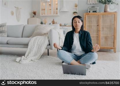 Girl sitting on floor in front of laptop in lotus pose and listening to tranquil music in airpods. Young european woman practicing yoga online at home for stress relief. Remote lesson on quarantine.. Girl sitting on floor in front of laptop in lotus pose and listening to tranquil music in airpods.