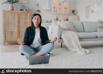 Girl sitting on floor in front of laptop in lotus pose and listening to tranquil music in airpods. Young european woman practicing yoga online at home for stress relief. Remote lesson on quarantine.. Girl sitting on floor in front of laptop in lotus pose and listening to tranquil music in airpods.
