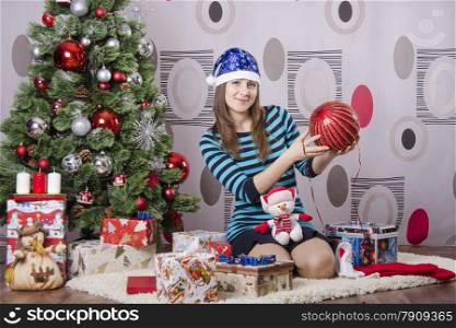 Girl sitting on the carpet beside the Christmas tree. In the hands of her big red Christmas ball.