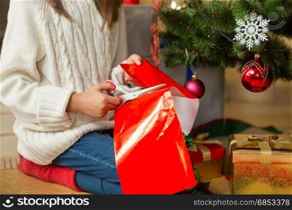 Girl sitting under Christmas tree and cutting red paper with scissors for decorating presents