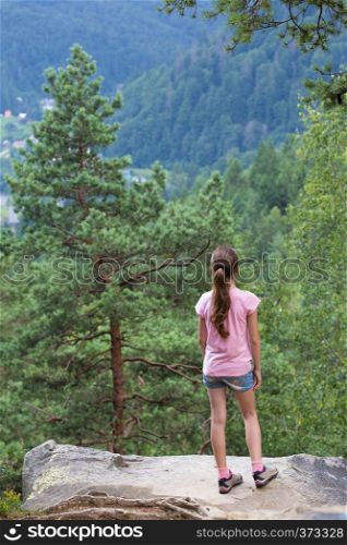 girl standing at the rocky cliff and looking at the forest mountains