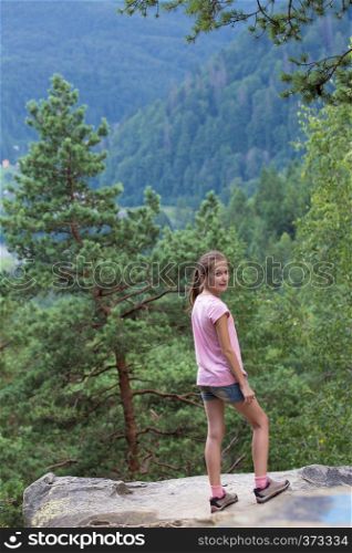 girl standing at the rocky cliff and looking at the forest mountains