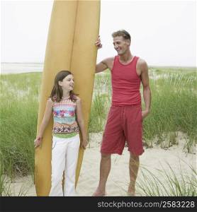 Girl standing in front of a surfboard beside her father and smiling
