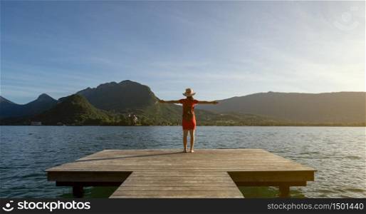 Girl standing up with a stalk hat on and red dress with her arms up looking at the mountains with her shoes on a dock by a lake. Lifestyle concept