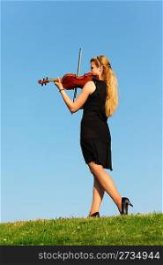 girl stands on grass and plays violin against sky