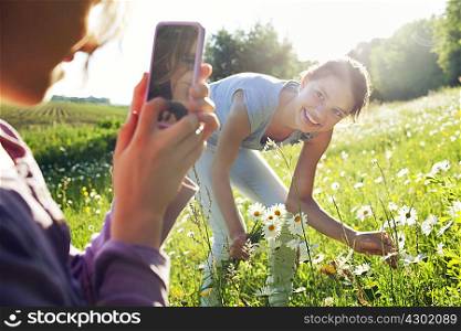 Girl taking photograph of girl picking flowers