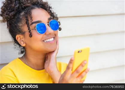 Girl teenager teen female young African American mixed race biracial woman outside smiling with perfect teeth wearing blue sunglasses and using a mobile cell phone for social media