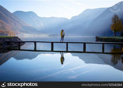 girl tourist in a hat and with a backpack stands on a wooden bridge on a mountain lake in the early morning. beautiful landscape and reflection