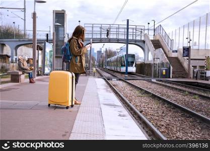 girl tourist with a backpack and a big yellow suitcase stands on the platform and waits for the train