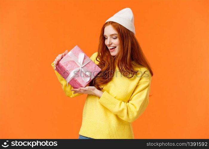 Girl trying figure-out whats inside gift box shaking it and looking intrigued. Excited happy redhead woman like winter holidays and receiving present, standing amused orange background.. Girl trying figure-out whats inside gift box shaking it and looking intrigued. Excited happy redhead woman like winter holidays and receiving present, standing amused orange background