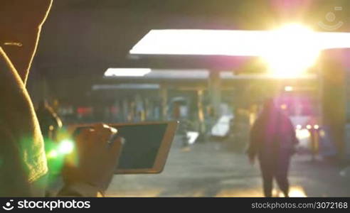 Girl typing on tablet computer at the station. Other people walking around. Bright evening sun shining in the sky