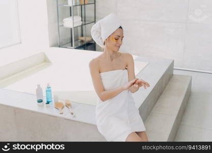 Girl uses eye patches   hand cream. Attractive woman in towel post-bath. Hispanic lady showers at home. Spa relaxation. Modern bathroom interior.