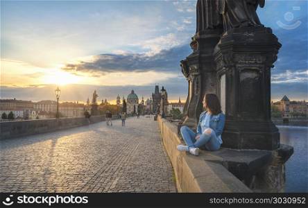 Girl watching the sunrise on Charles Bridge. Young brunette woman, sitting relaxed on the Charles Bridge walls, over the Vltava river, admiring the sunrise over Prague City, in Czech Republic.
