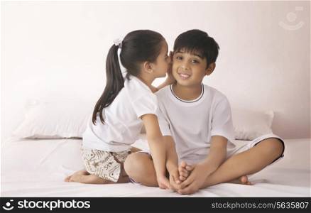 Girl whispering in brother&rsquo;s ear on bed