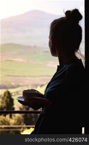 Girl with a cup of coffee is standing by the window outside the window Tuscan landscape. Tuscany, Italy