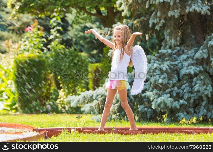 Girl with angel wings in the garden enjoys splashes of water. Happy six year old girl dressed as an angel with wings is walking with splashes of water for watering the lawn