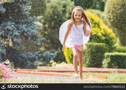 Girl with angel wings running around in the rain in the garden. Happy six year old girl dressed as an angel with wings is walking with splashes of water for watering the lawn
